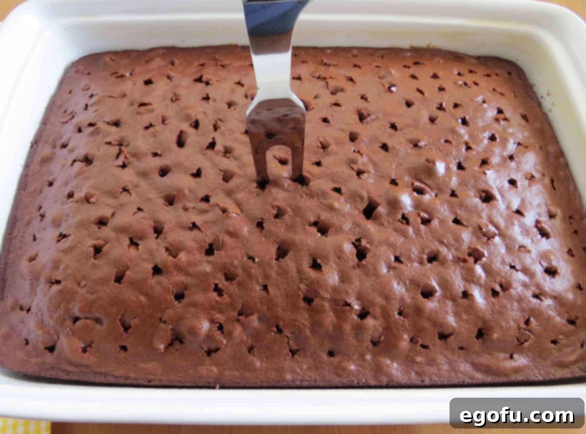 a large meat fork piercing holes in fully baked chocolate cake in a white baking dish.