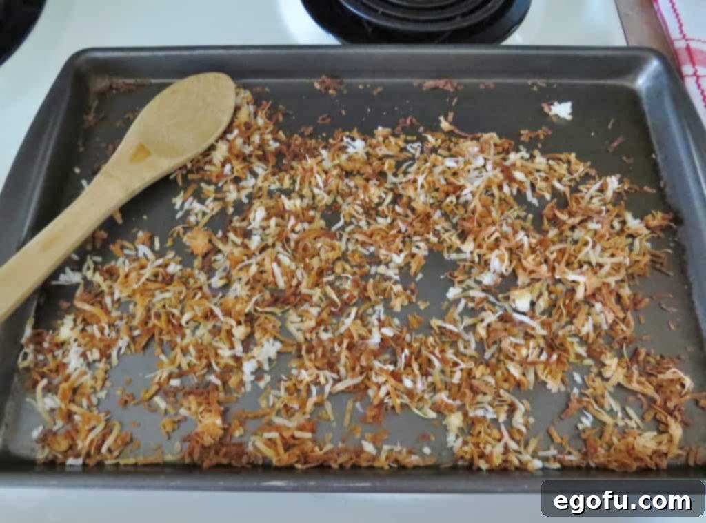 Toasted coconut, now a rich golden-brown and fragrant, spread on a cookie sheet next to a wooden spoon, indicating it's perfectly done and ready to cool before garnishing.