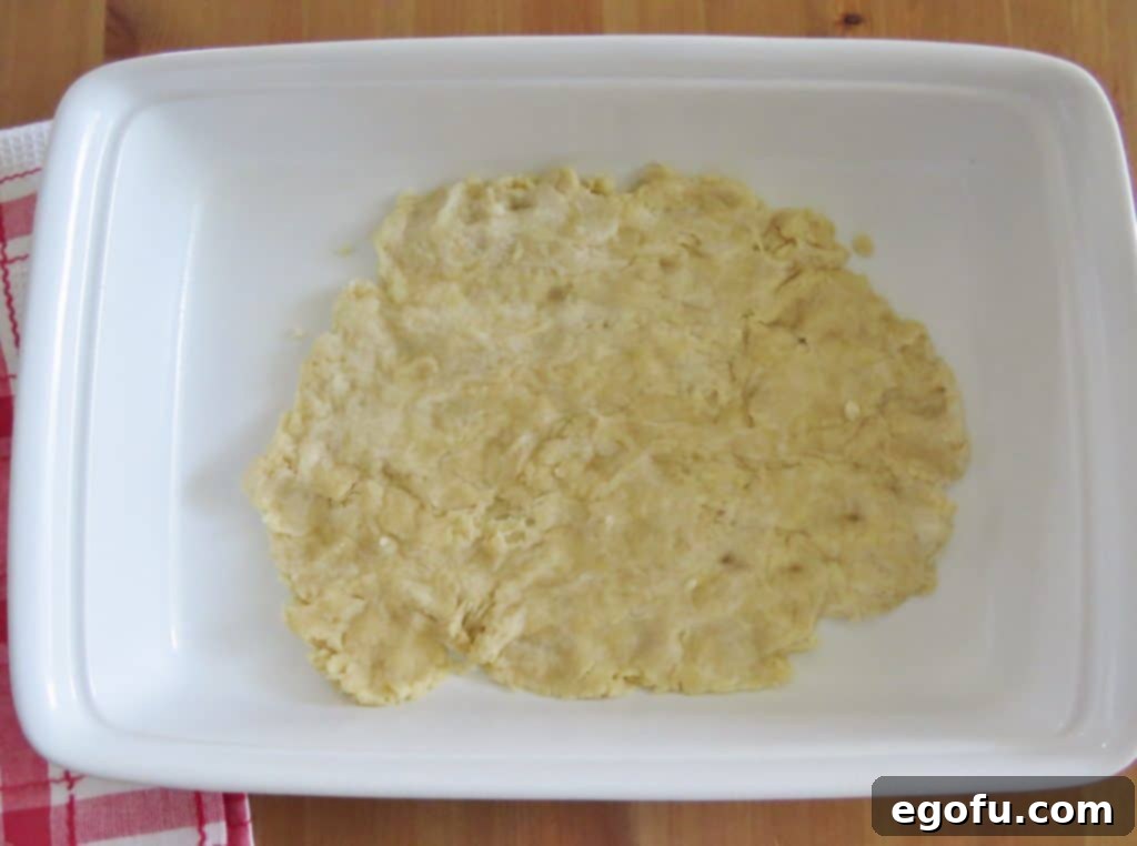 Homemade pie crust dough being carefully spread and pressed into the bottom of a white rectangular baking dish, ready for its initial bake.