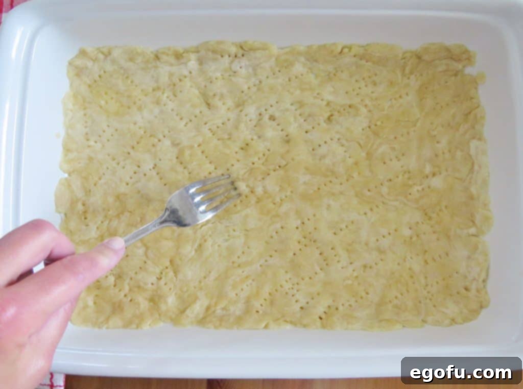 Close-up view of a pie crust in a baking dish, with a fork making numerous indentations across its surface to prevent steam bubbles during baking.
