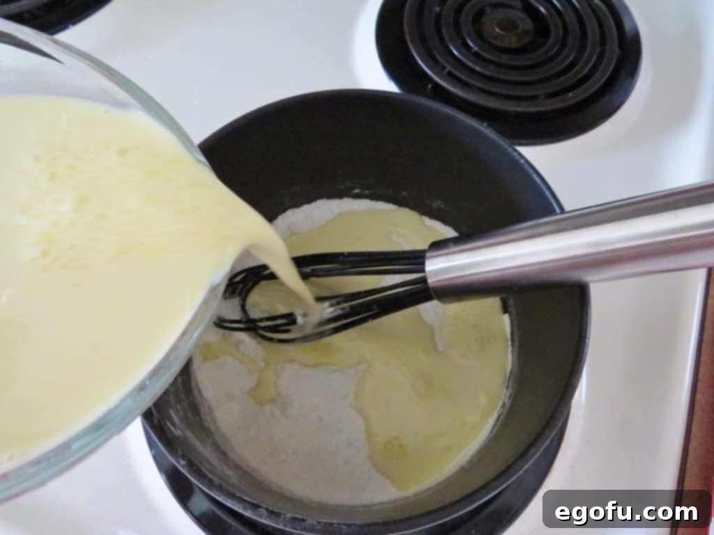 A stream of half and half and egg yolk mixture being slowly poured into a saucepan containing sugar and cornstarch, with a whisk prepared to combine the ingredients smoothly.