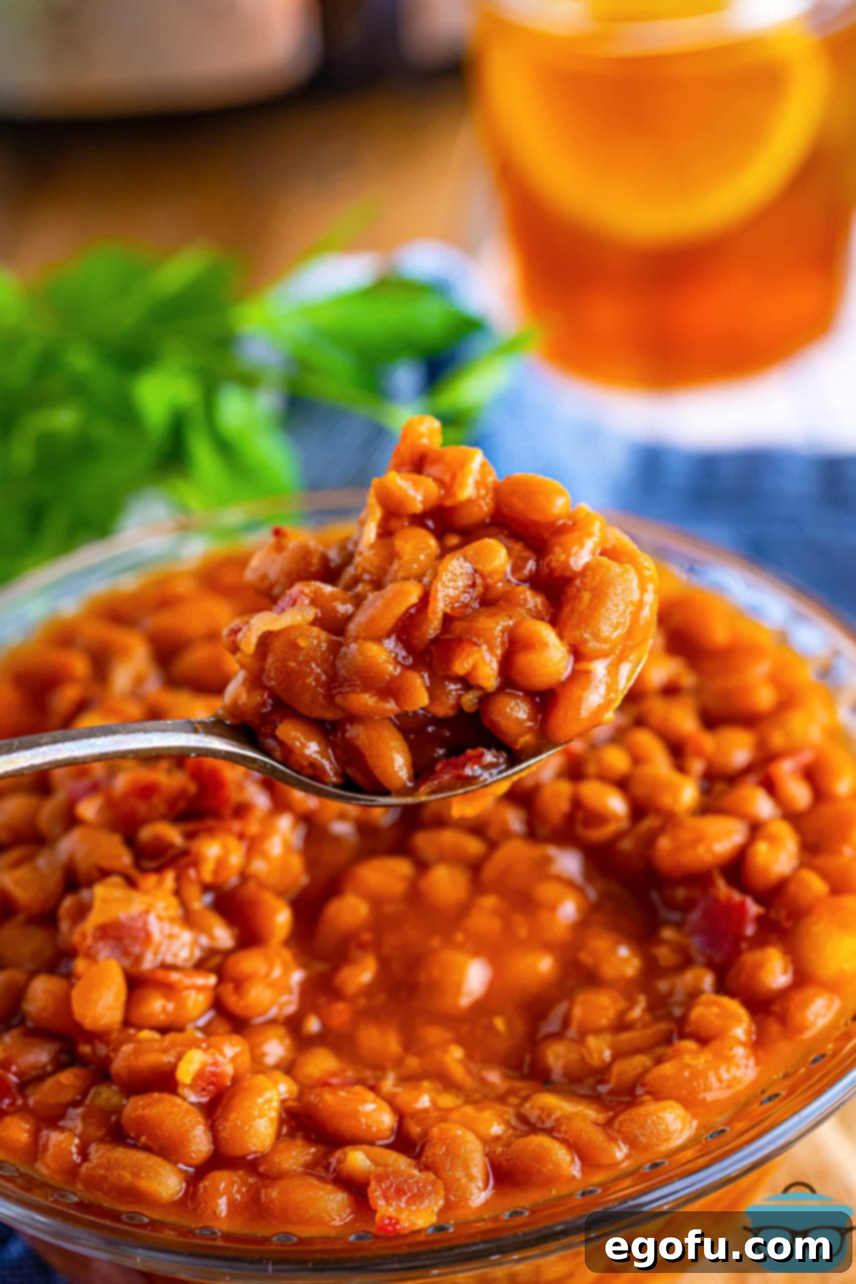 A large serving bowl overflowing with glistening Crock Pot Baked Beans, topped with crispy bacon bits, with a serving spoon poised for a generous scoop.
