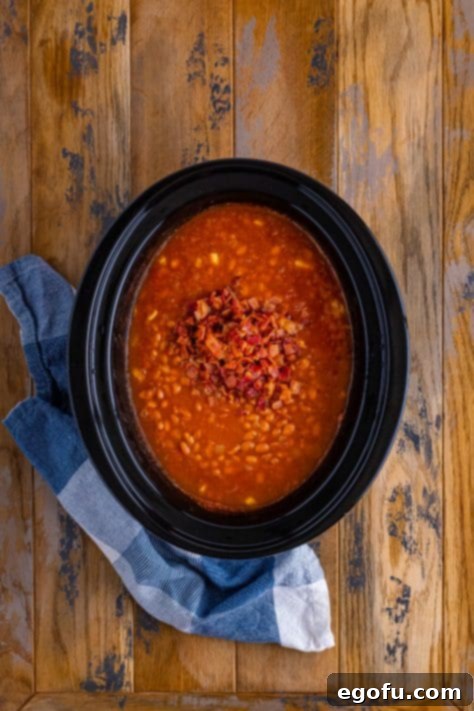 Crispy, crumbled bacon being gently incorporated into the hot, freshly cooked Crock Pot Baked Beans within the slow cooker insert.