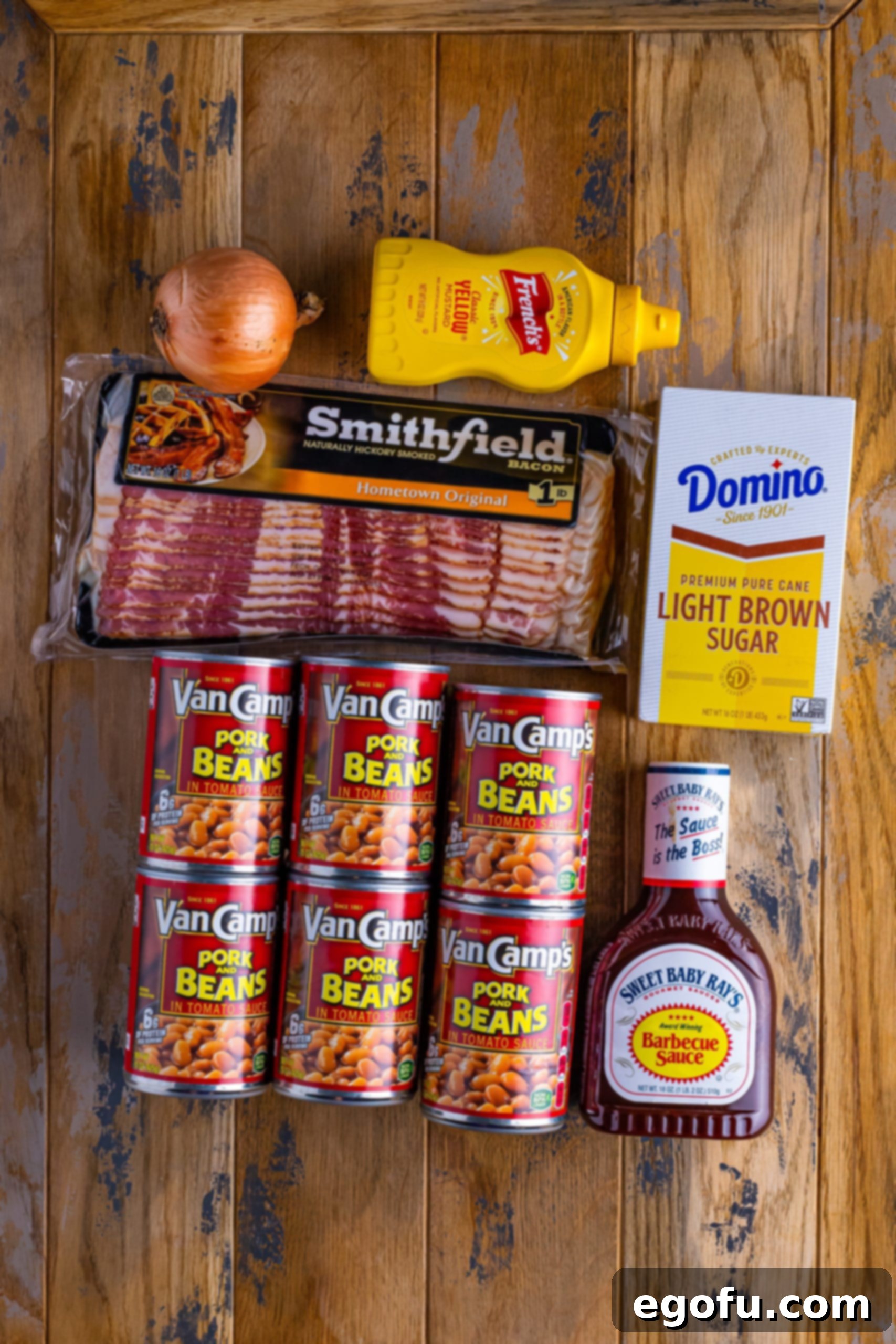 A flat lay photograph displaying all the essential ingredients for slow cooker baked beans: canned pork and beans, freshly diced onion, a bottle of barbecue sauce, a container of yellow mustard, a bag of light brown sugar, and a bowl of crispy cooked bacon.