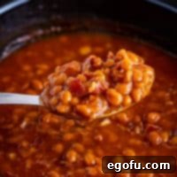 A scoop of rich, saucy Crock Pot Baked Beans, garnished with crispy bacon, being lifted by a spoon over the slow cooker.