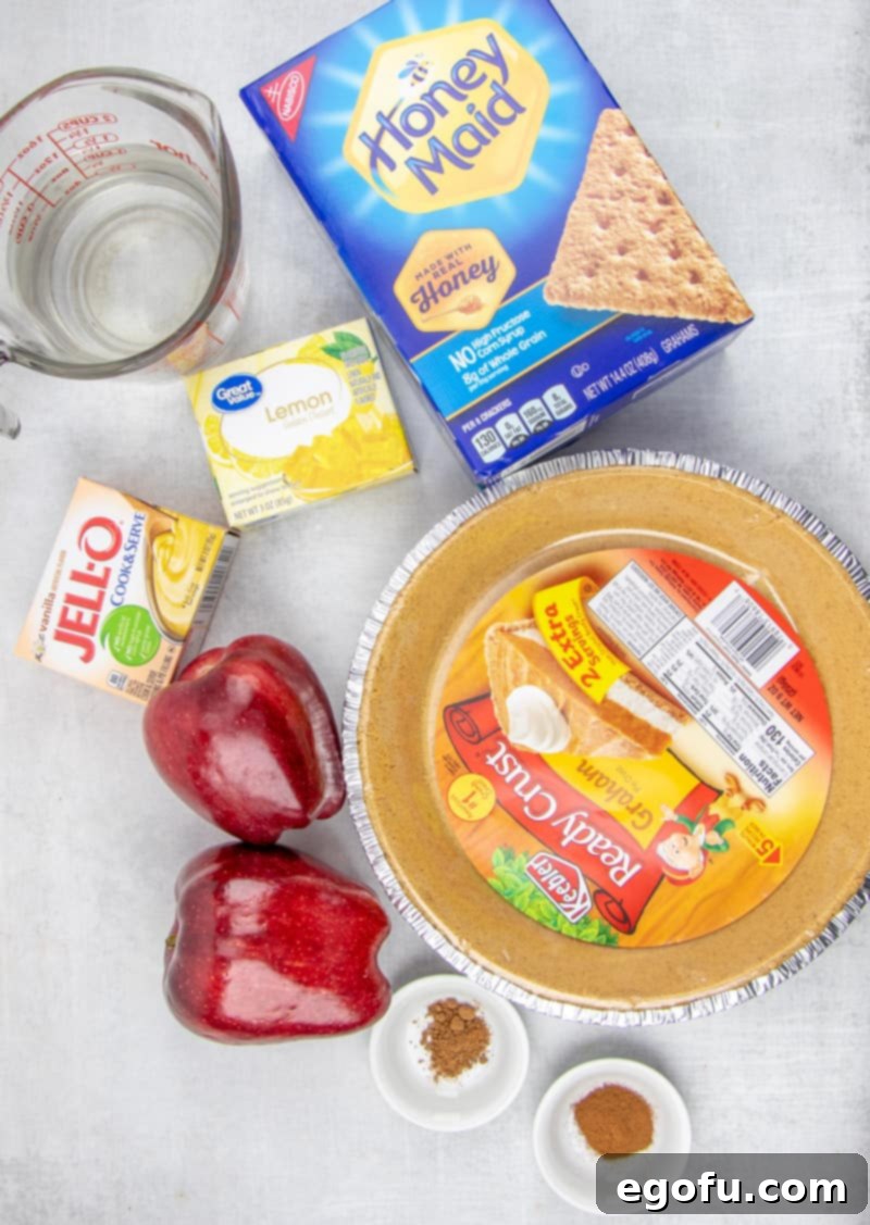 All ingredients for a no-bake apple pie laid out on a wooden surface: graham cracker crust, neatly sliced apples, a box of lemon Jell-O, vanilla pudding mix, a glass of water, cinnamon, and nutmeg, highlighting the simplicity of this quick dessert.