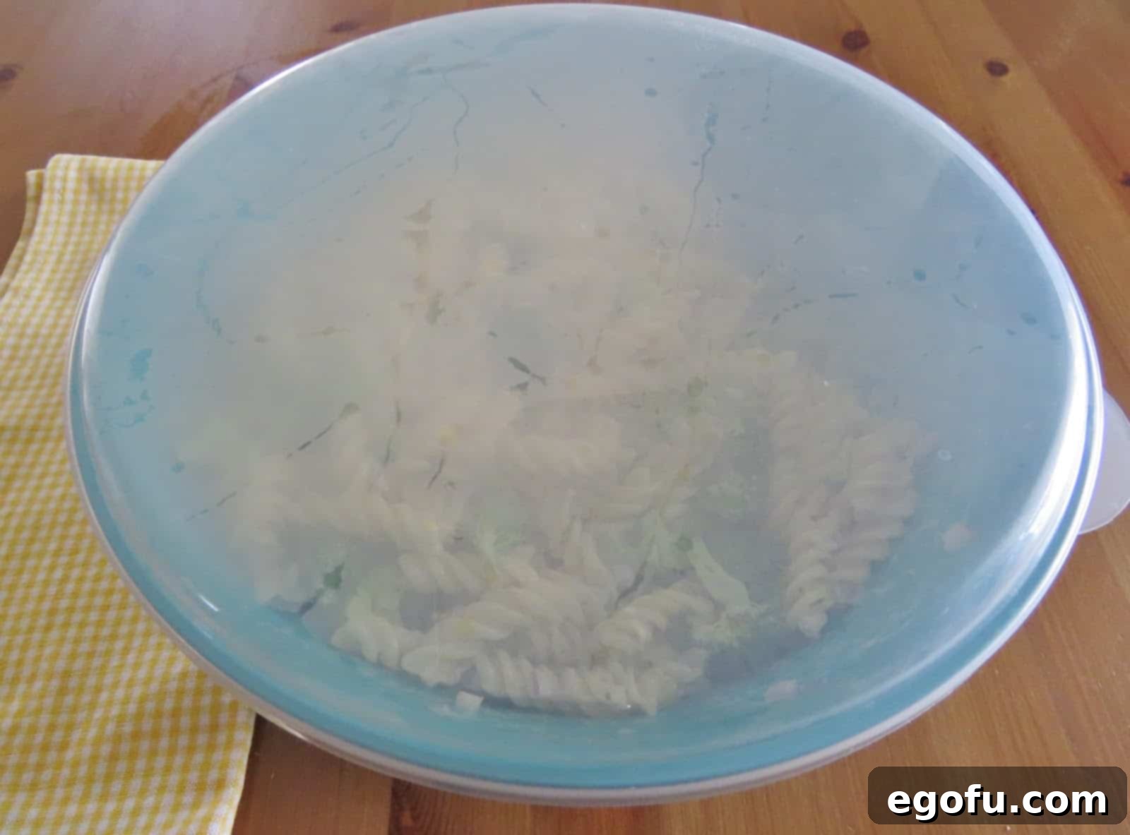 A large plastic blue bowl filled with the uncooked pasta salad mixture (pasta, broccoli, red onion, dressing), covered and ready for chilling in the refrigerator.