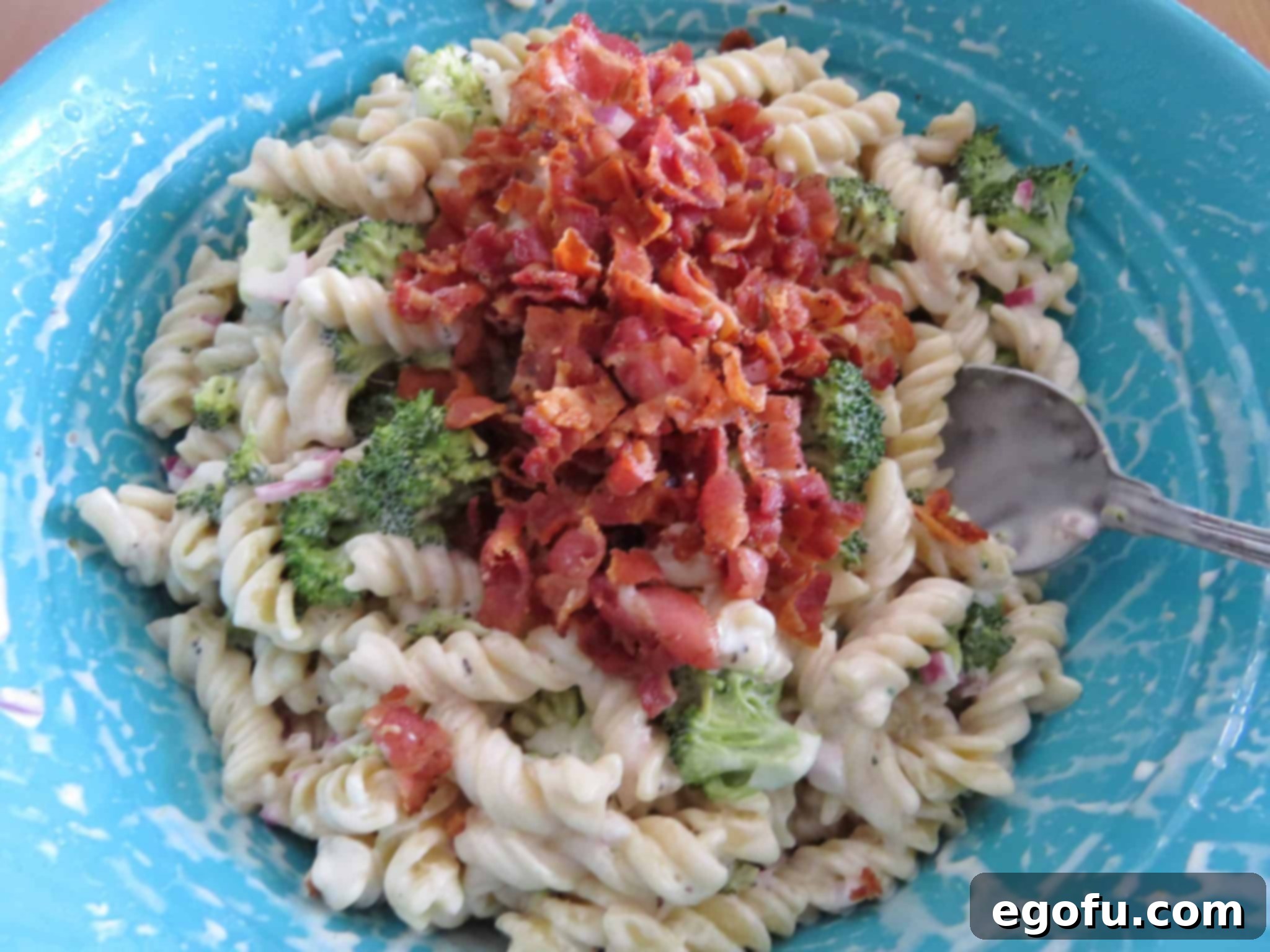 Cooked crumbled bacon being added to the chilled pasta salad with broccoli florets, diced red onion, and rotini pasta, all mixed in a creamy dressing in a blue bowl.