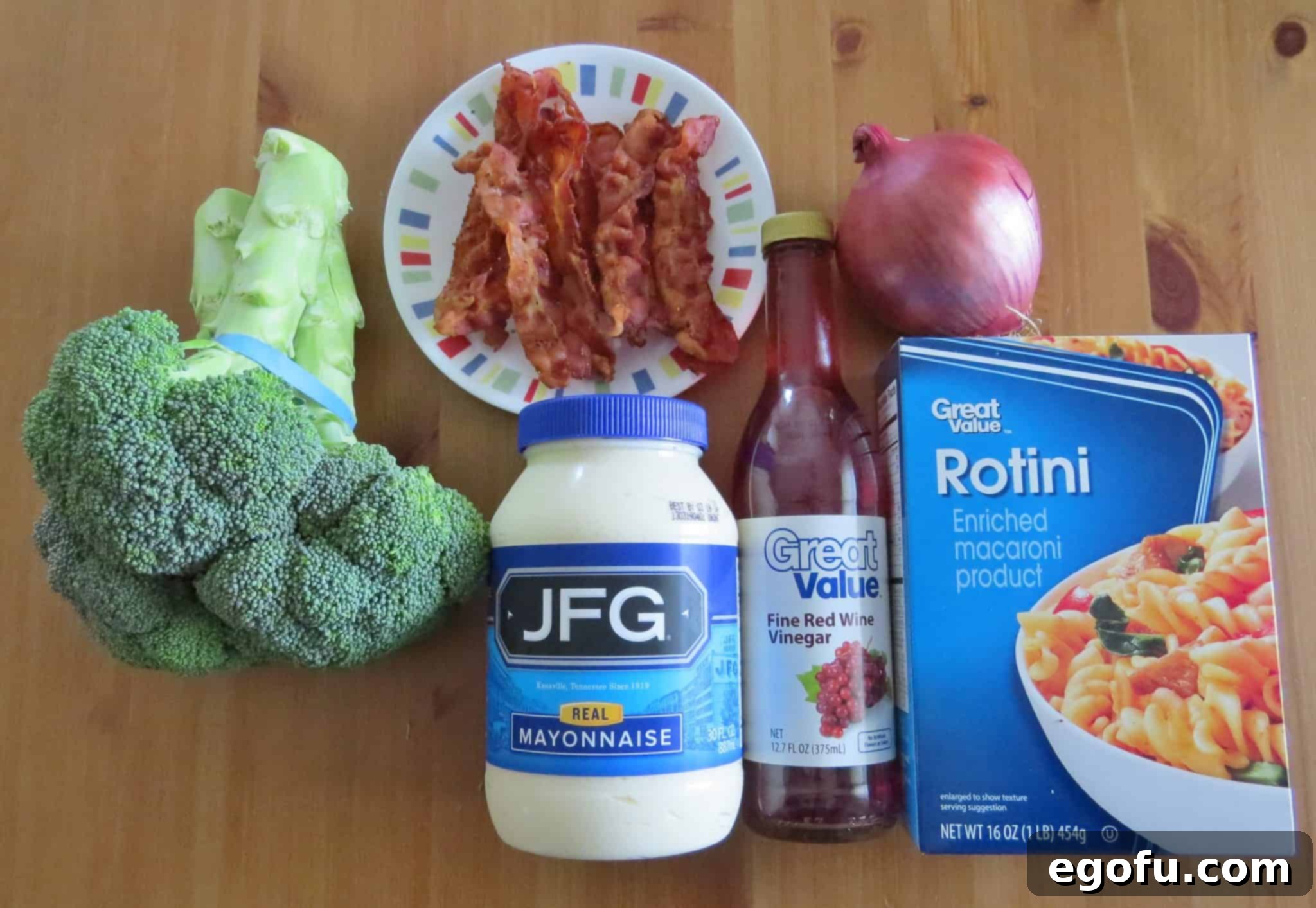 An array of fresh ingredients: rotini pasta, fresh broccoli, red onion, bacon, mayonnaise, red wine vinegar, sugar, salt, and pepper, laid out on a wooden surface.