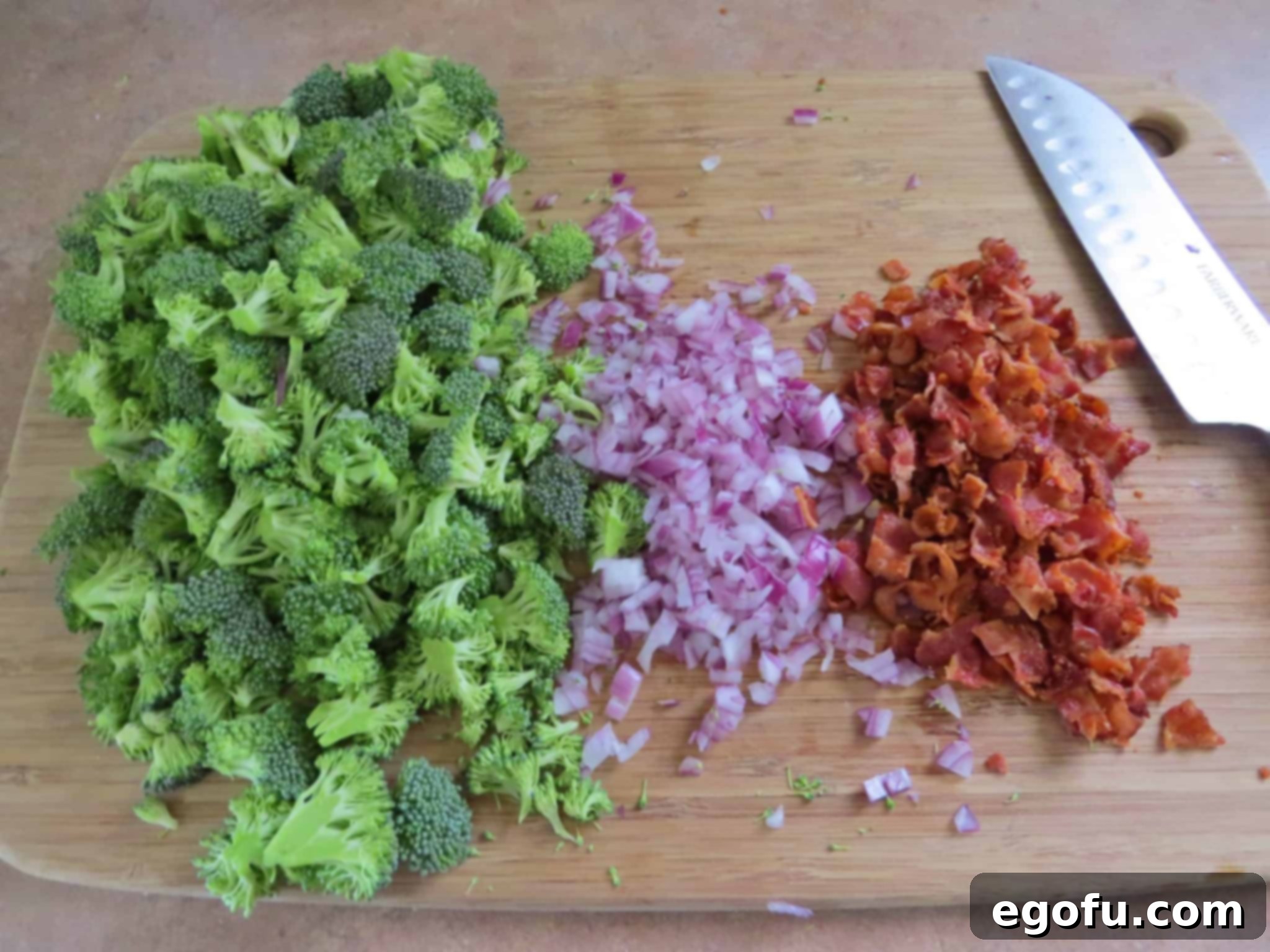 A collection of prepped ingredients: finely chopped broccoli florets, diced red onion, and crumbled cooked bacon, arranged on a wooden chopping board with a knife.