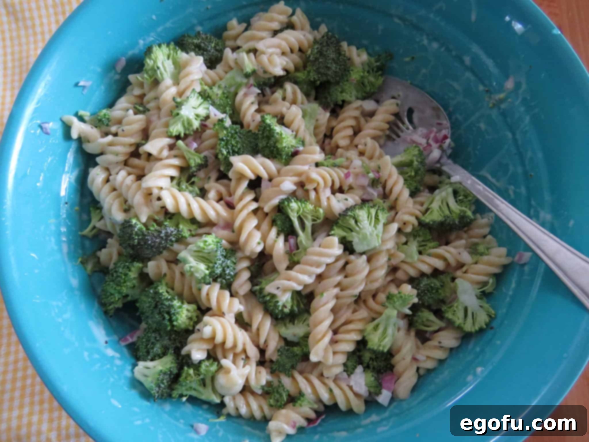 The pasta salad ingredients, including rotini, broccoli, and red onion, being mixed together with the creamy dressing in a large blue bowl.