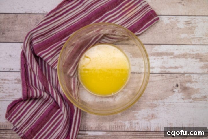 A pristine clear glass bowl containing a pool of perfectly melted unsalted butter, ready for dipping and coating, sitting on a clean white surface.