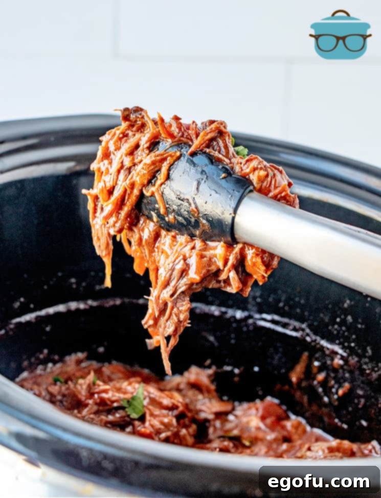 A pair of tongs lifting a generous portion of Mexican-Style Shredded Beef directly from the slow cooker, showcasing its rich texture and readiness to serve.
