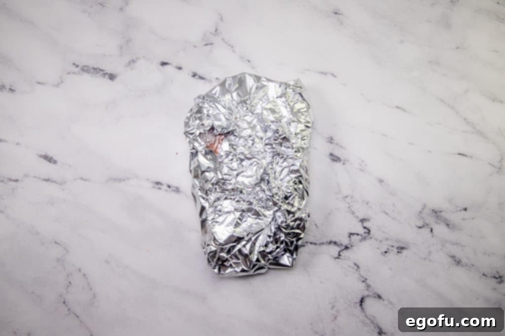 Fiesta-Ready Slow Cooker Shredded Beef 7 A seasoned chuck roast, now wrapped in both plastic wrap and aluminum foil, resting on a marble surface, prepared for refrigeration.