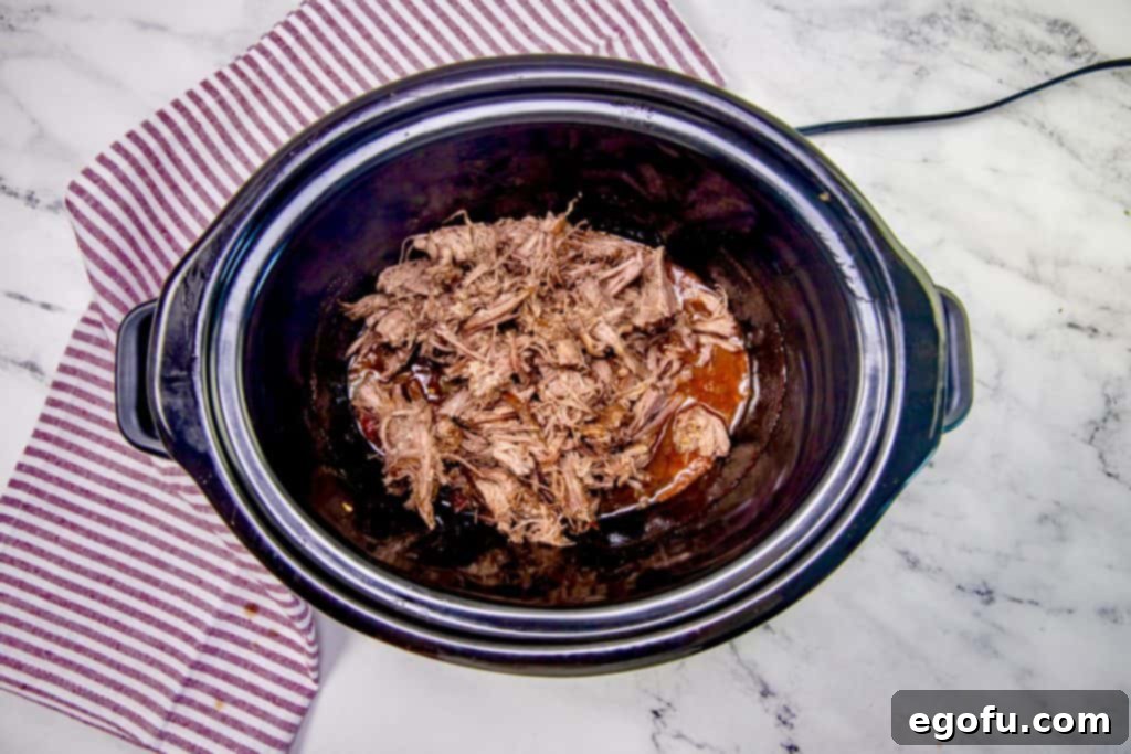 Perfectly cooked and shredded chuck roast, steaming gently, inside a black oval slow cooker, ready for its final flavoring.