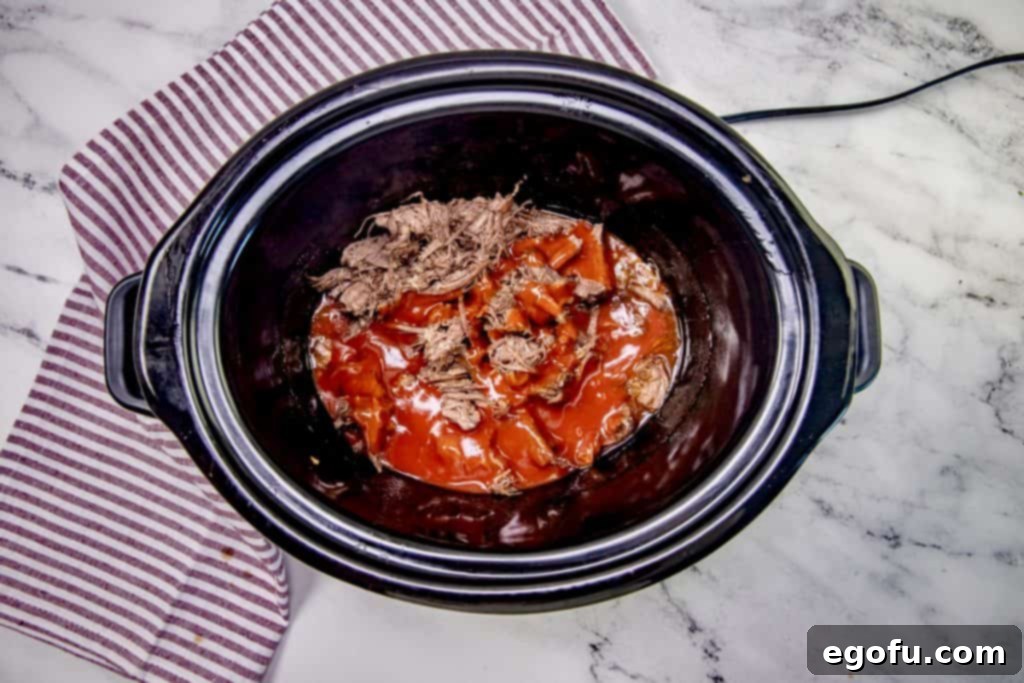 Taco sauce being poured over the shredded and cooked chuck roast in the slow cooker, ready to be stirred and heated for final flavor infusion.