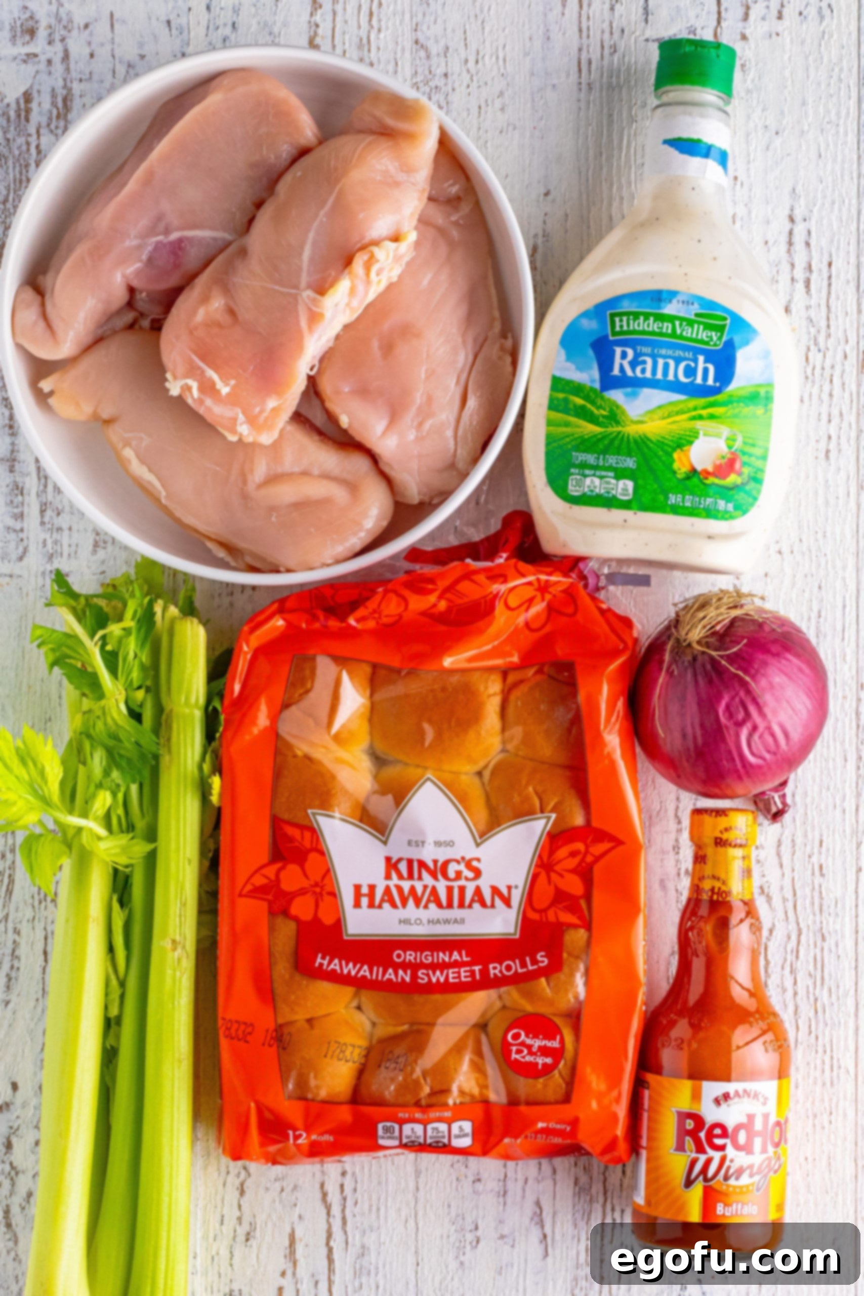 A flat lay of all the essential ingredients: Hawaiian slider rolls, a bottle of ranch dressing, chopped celery, raw chicken breasts, a bottle of buffalo hot sauce, and a red onion.