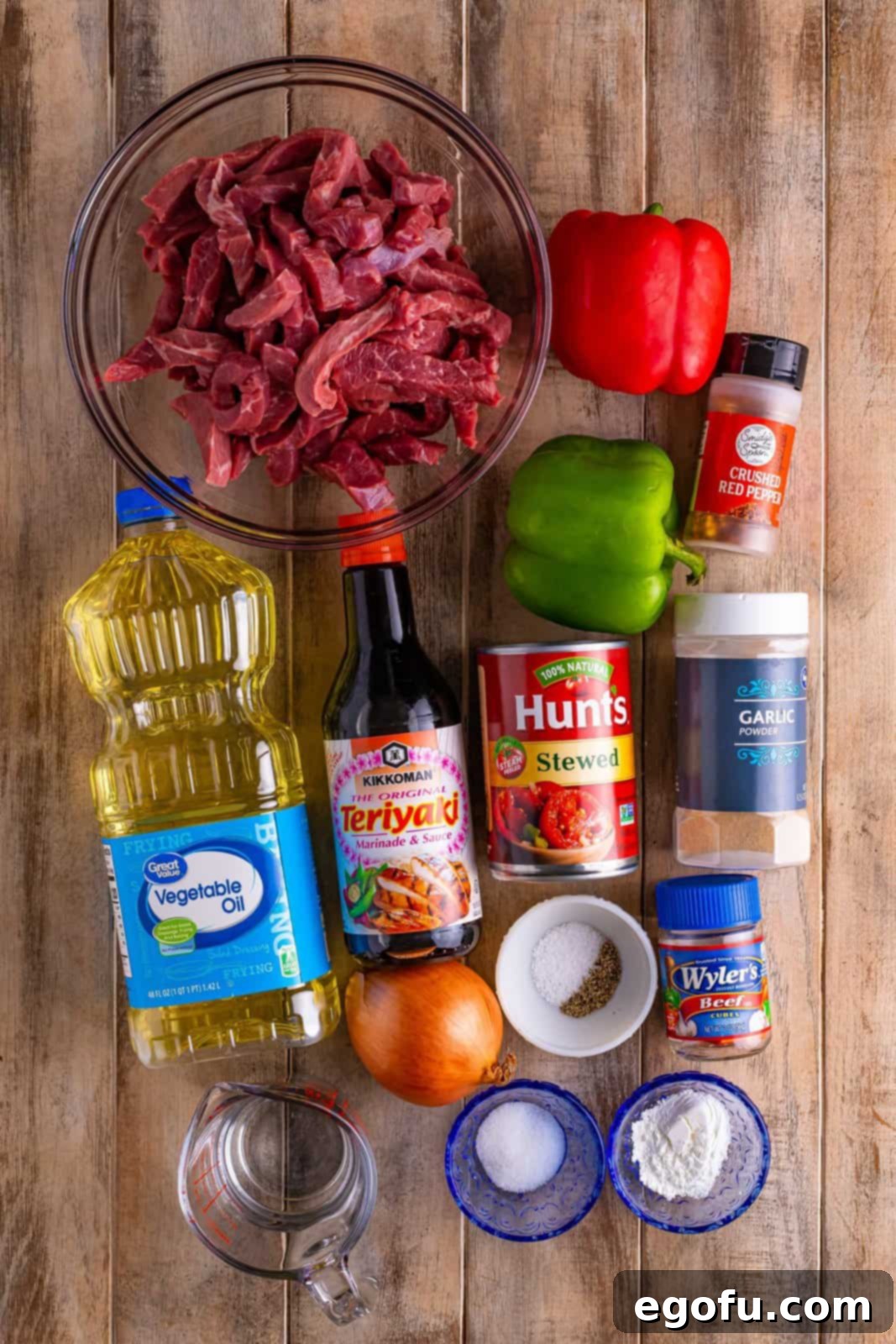 Slow Cooker Pepper Steak 4 A flat lay showing uncooked fajita beef strips, canned stewed tomatoes, sliced red and green bell peppers, onion, garlic powder, red pepper flakes, beef broth, and soy sauce – all ingredients for Crock Pot Pepper Steak.