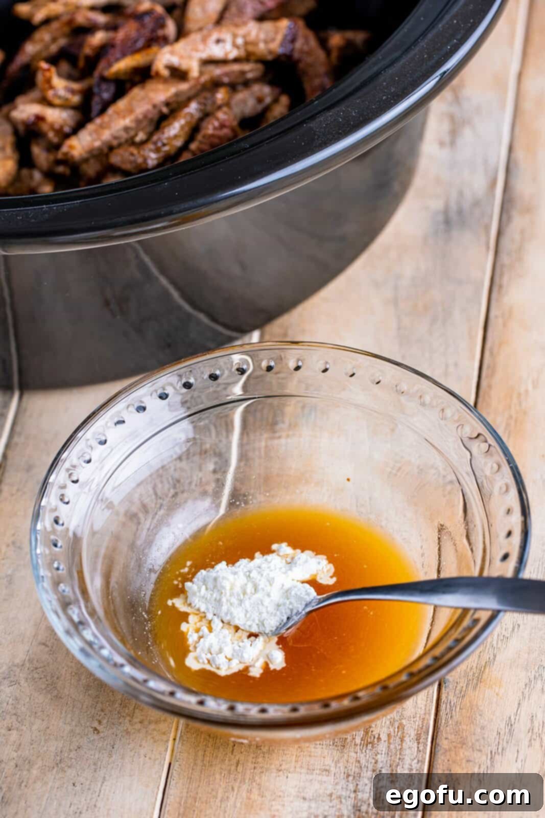 Slow Cooker Pepper Steak 9 Cornstarch added to the beef bouillon and water mixture in a small clear bowl, being stirred with a fork to form a slurry.