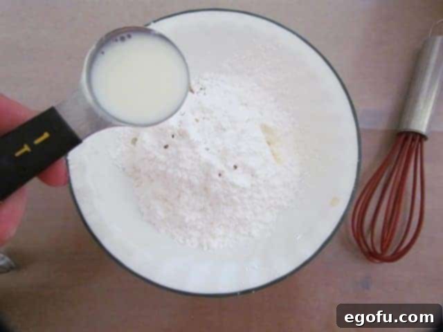 milk being poured into a white bowl containing powdered sugar and clear vanilla extract