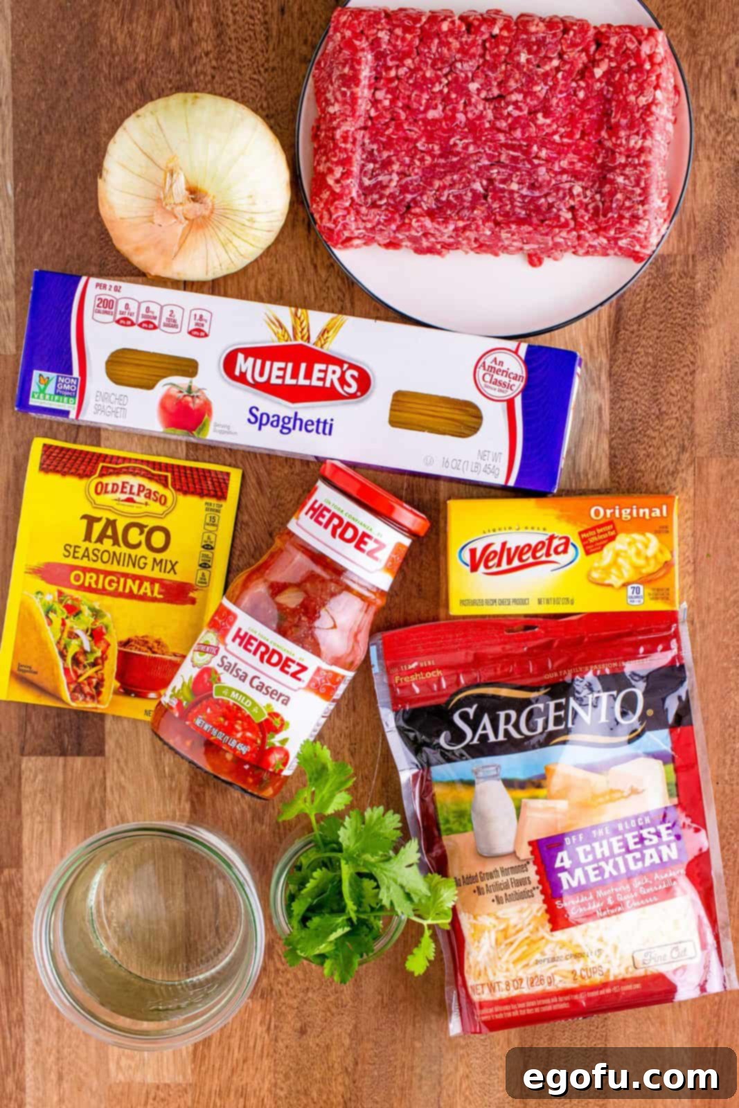 All the necessary ingredients laid out for One Pot Taco Spaghetti: ground beef, onion, taco seasoning, salsa, spaghetti, water, velveeta cheese, mexican shredded cheese, and fresh cilantro.