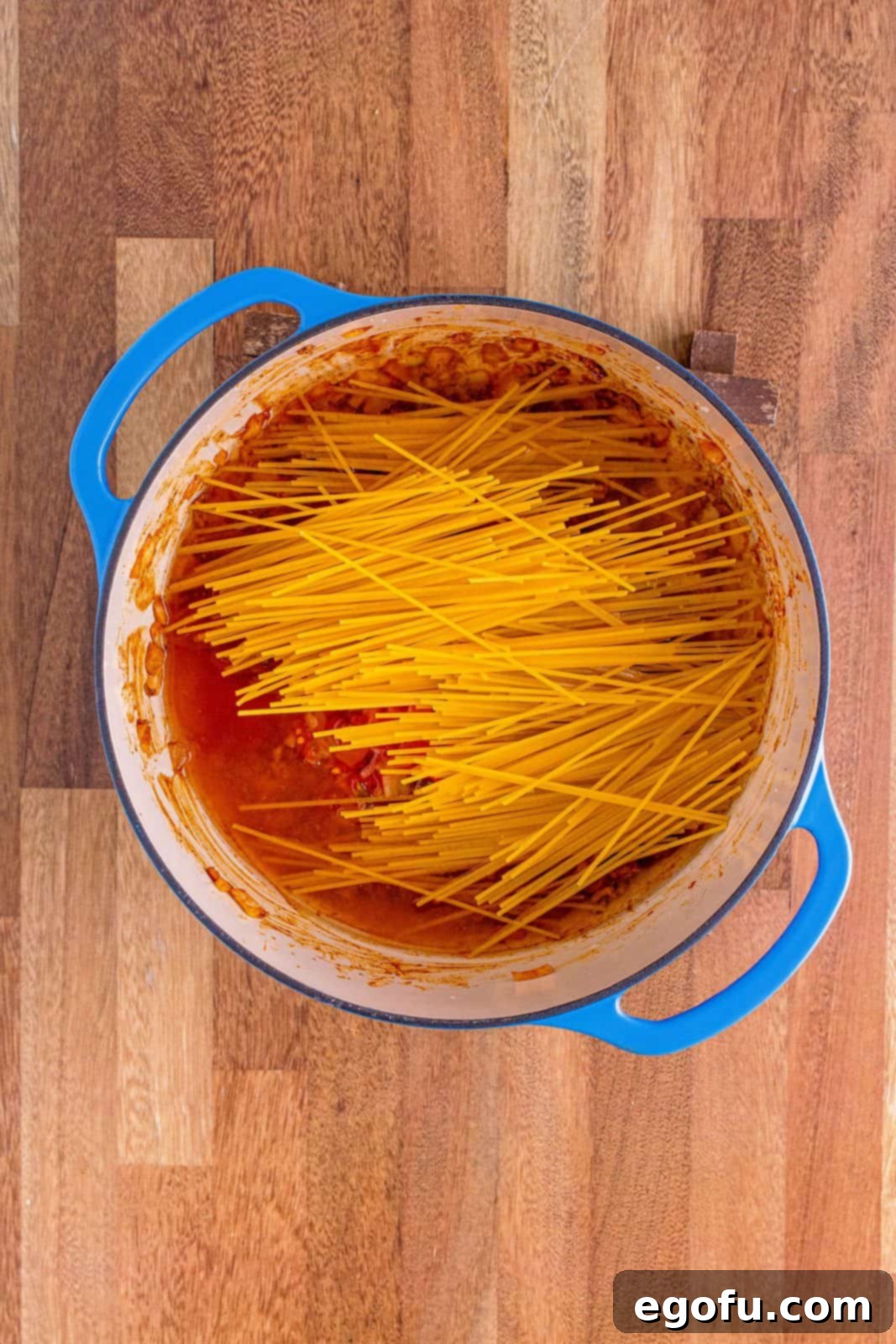 Salsa, broken spaghetti, and water are poured into the ground beef and onion mixture in the Dutch oven, preparing for cooking.