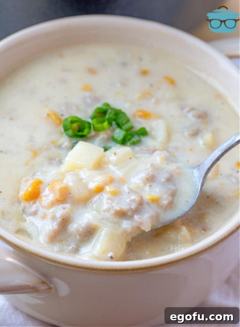 A white bowl filled with creamy sausage corn chowder, garnished with fresh green onions, with a spoon scooping up a delicious portion.