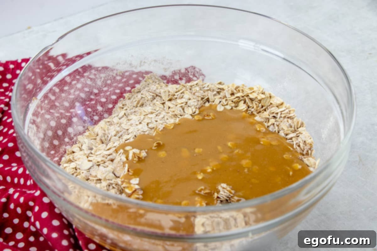 The peanut butter and honey mixture being poured over the oat mixture in a clear bowl, ready for combining.