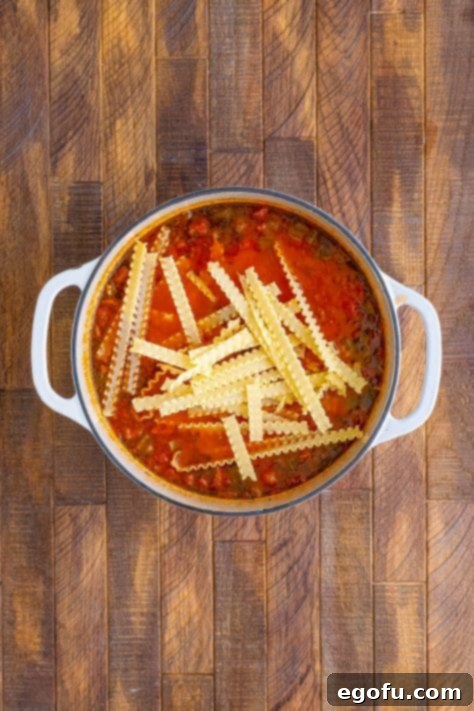 Mafalda pasta being added to the simmering lasagna soup in a large pot.