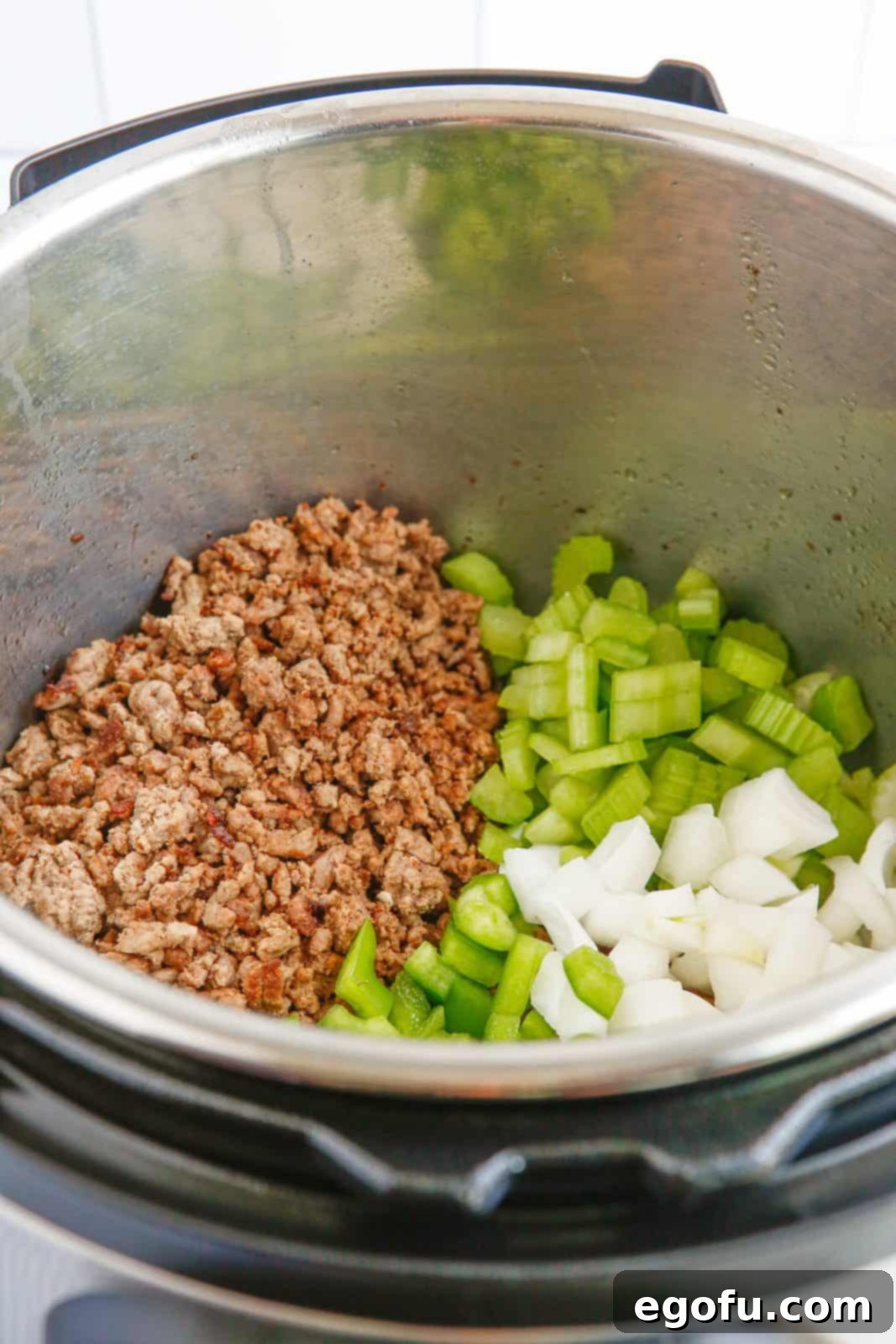 Diced yellow onions, celery, and green bell peppers being added to browned and crumbled ground beef in the Instant Pot, initiating the sautéing process for the aromatic 'holy trinity'.