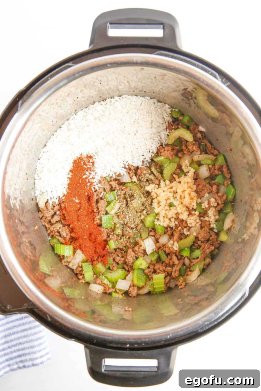 All the ingredients for Instant Pot Dirty Rice, including uncooked long grain rice, beef broth, a medley of spices, and a bay leaf, carefully layered in the Instant Pot, waiting to be sealed and pressure cooked.