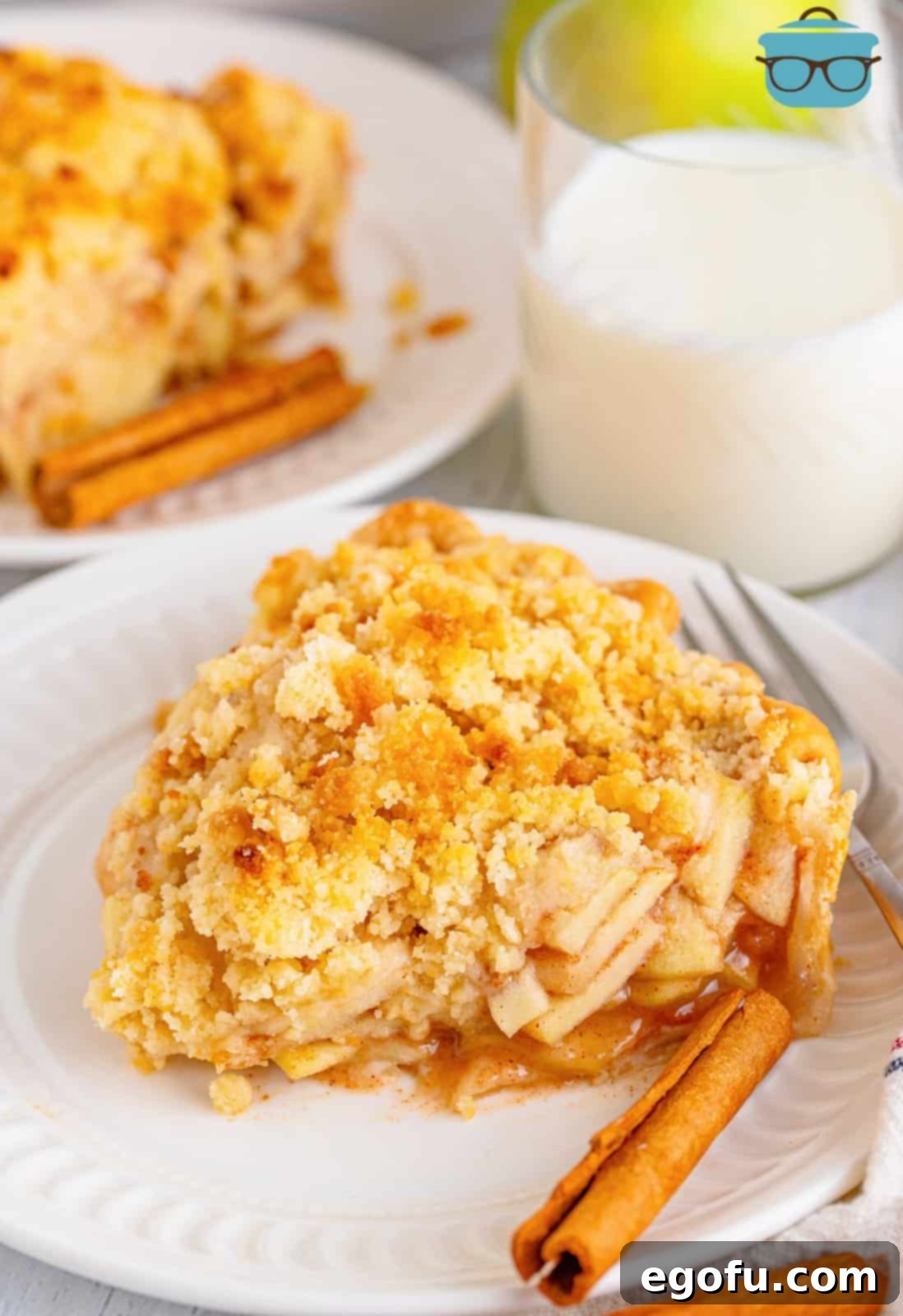 A perfectly sliced butter crumble apple pie on a white plate, with a glass of milk in the background, showcasing its golden crumble topping and rich apple filling.
