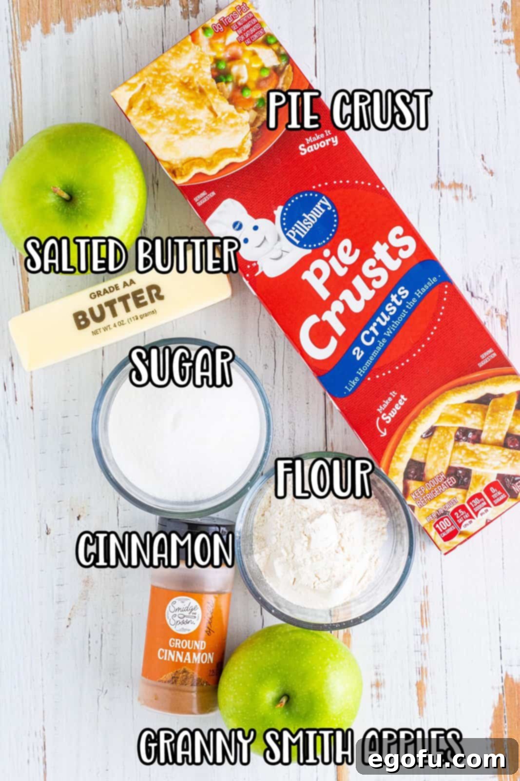 A flat lay of all the ingredients needed for butter crumble apple pie: refrigerated pie crust, fresh Granny Smith apples, a stick of salted butter, granulated sugar, all-purpose flour, and ground cinnamon.