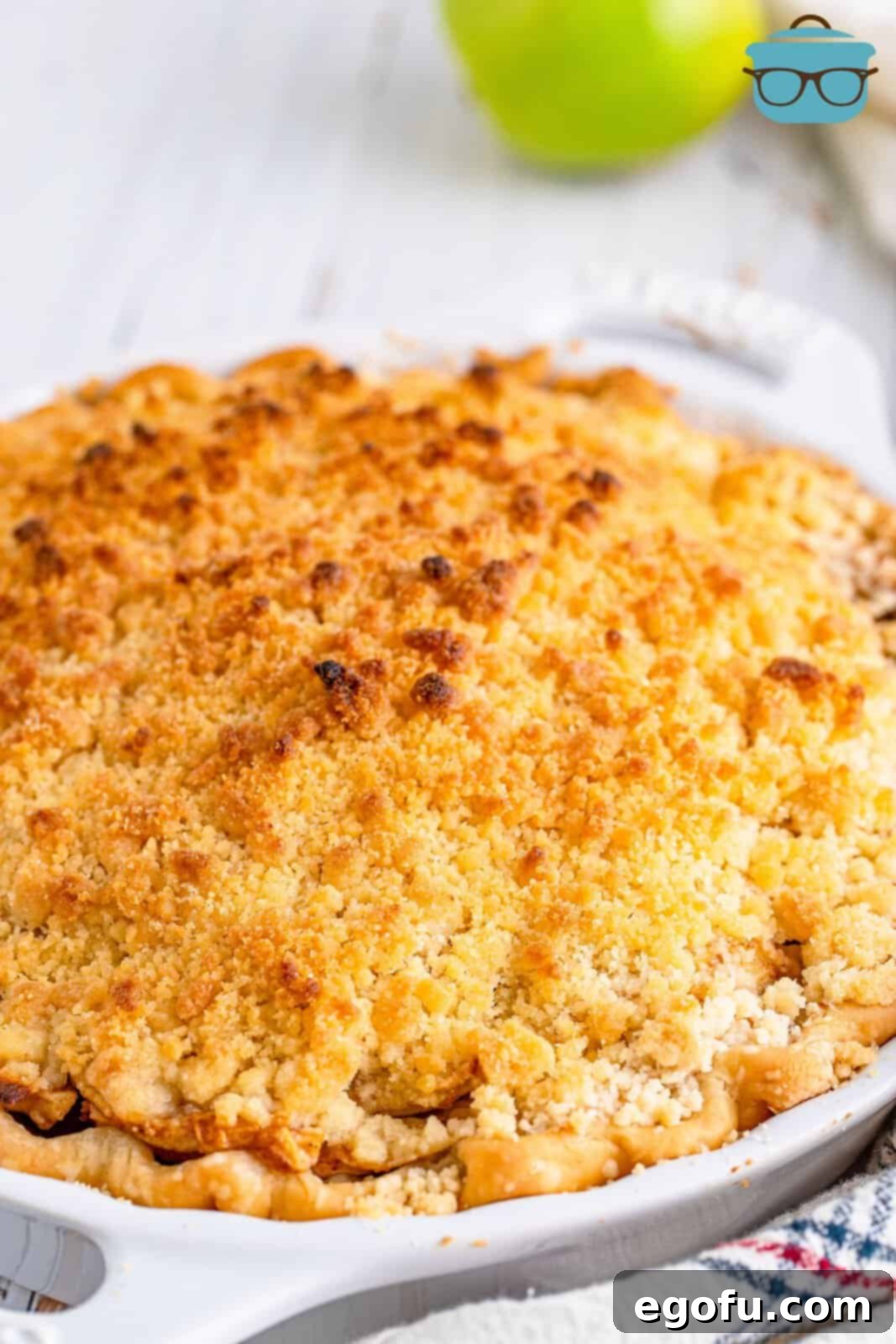 A full, beautifully baked butter crumble apple pie in a white ceramic pie pan, showcasing its golden-brown topping and perfectly set crust.