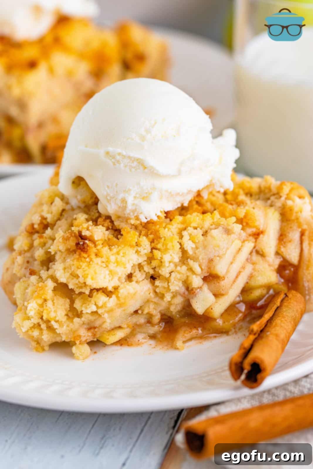A close-up photo of a single slice of butter crumble apple pie on a white plate, topped with a melting scoop of vanilla ice cream, highlighting the texture of the crumble and the tender apple filling.