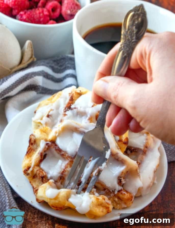 cinnamon roll waffles shown on a small white plate with a fork taking out a piece of the waffle.