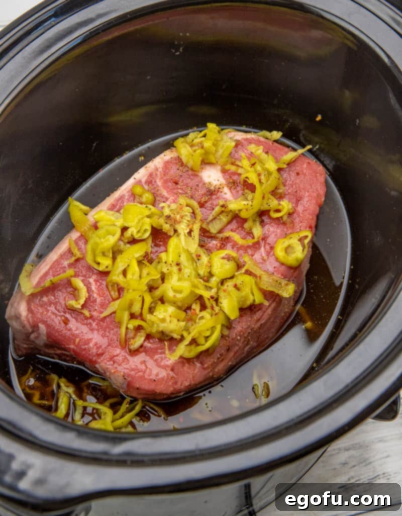 Chuck roast, beef broth, Italian dressing seasoning, and peperoncini simmering together in an oval slow cooker.