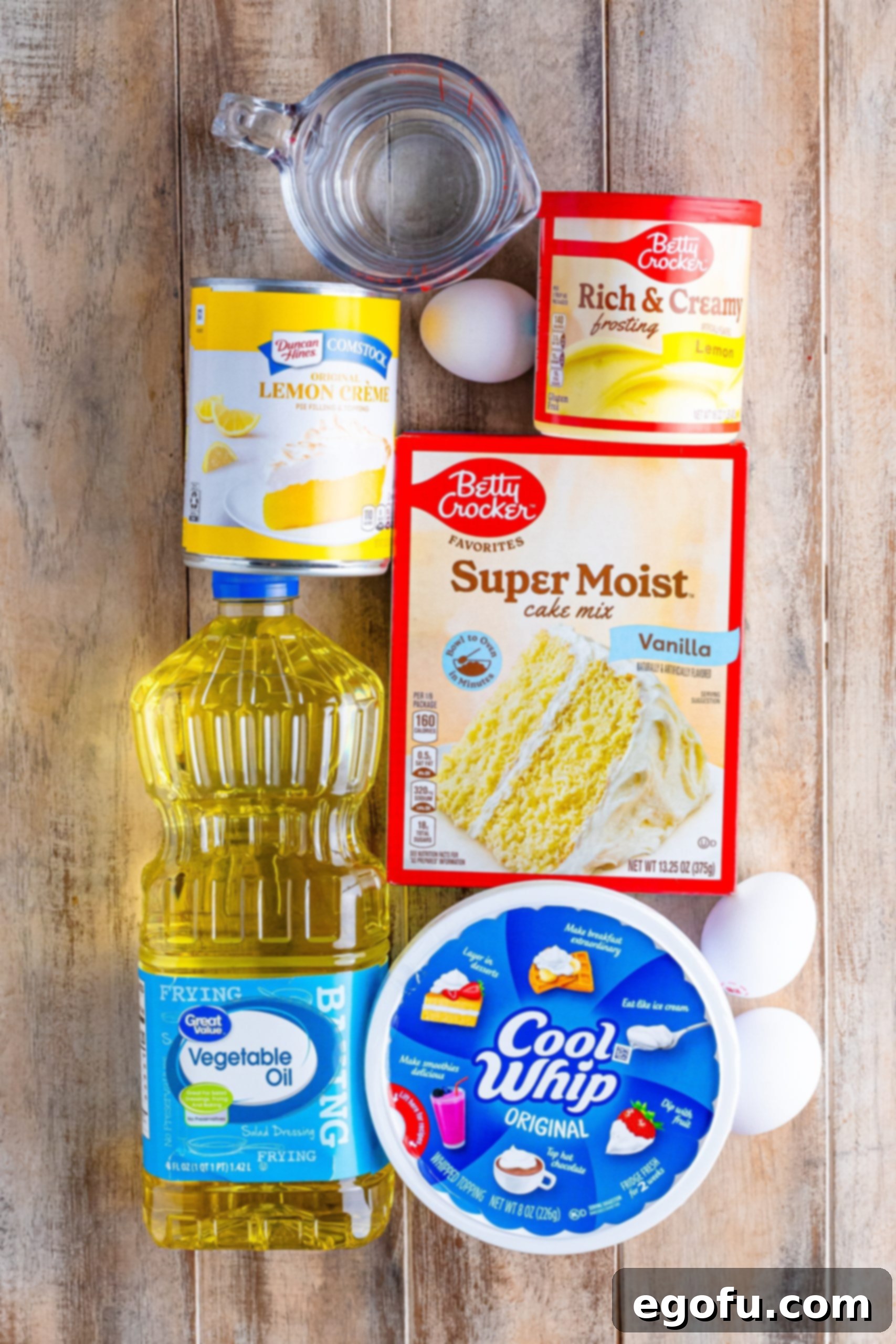 A neatly arranged flat lay showcasing all the essential ingredients for making Lemon Dream Cake: a tub of Cool Whip, a bottle of vegetable oil, fresh eggs, a glass of water, a tub of lemon frosting, a can of lemon pie filling, and a box of vanilla cake mix.