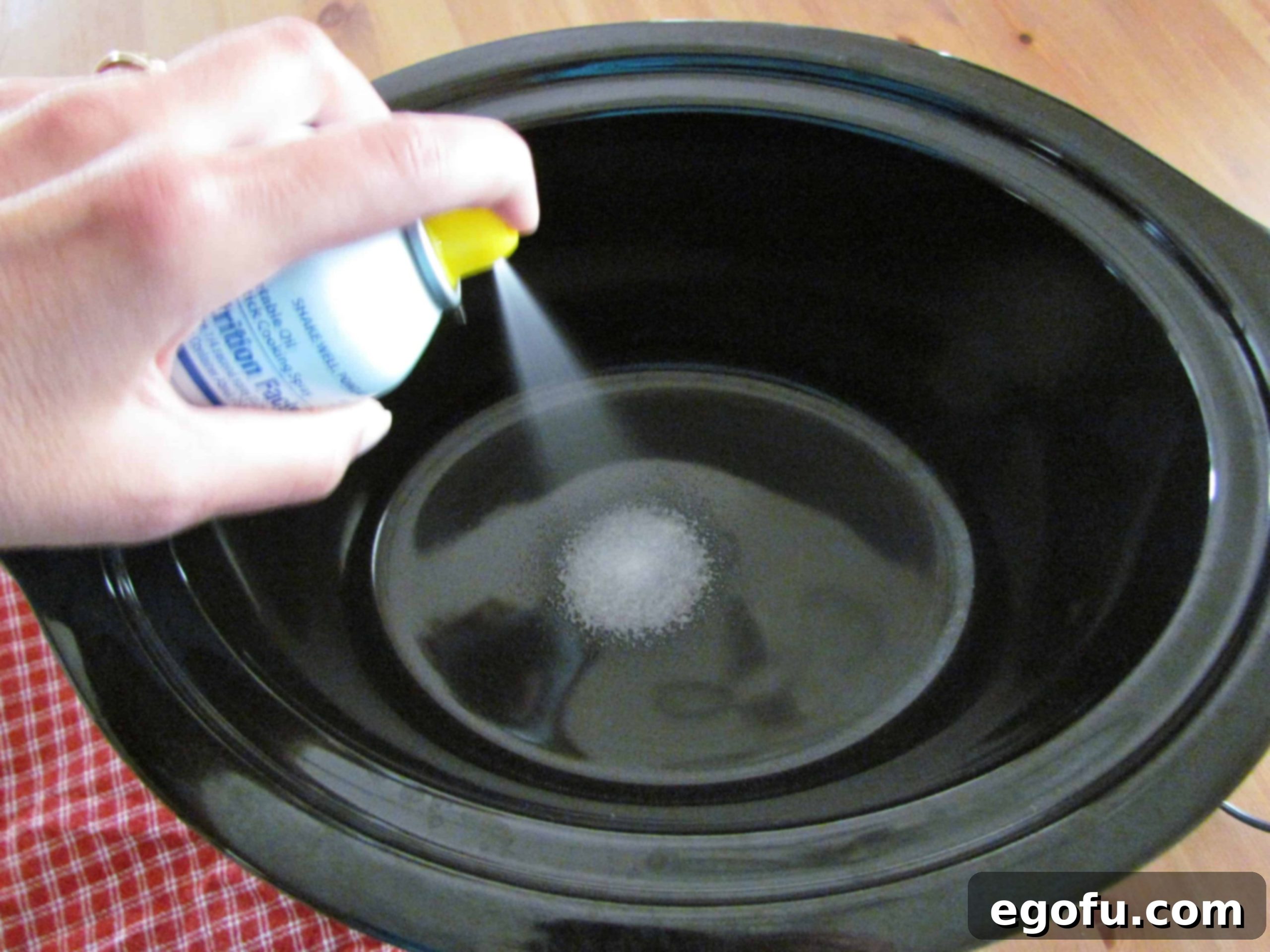 A hand spraying a black oval slow cooker with nonstick cooking spray, preparing it for the casserole.