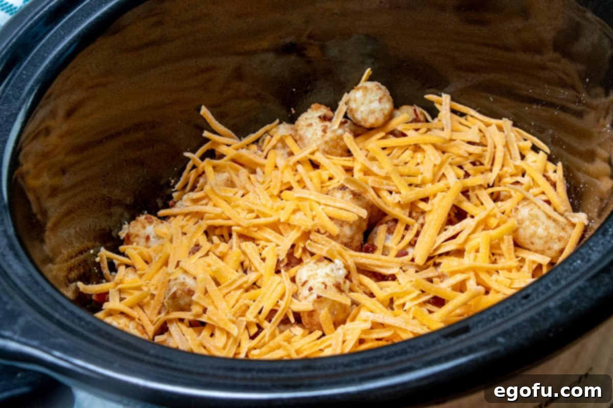 Shredded cheddar cheese now covers the bacon and tater tot layers in the slow cooker, creating a savory foundation.
