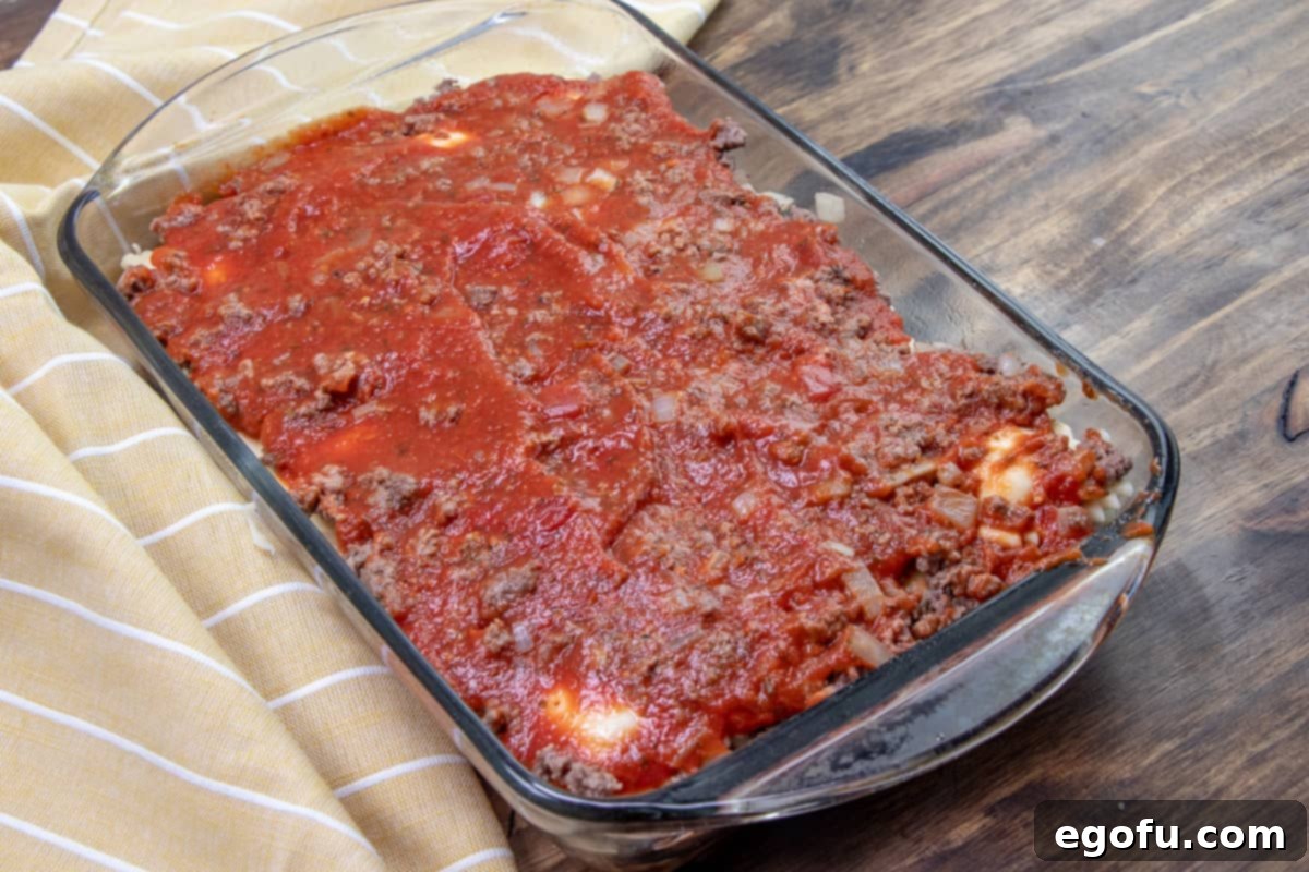 Another layer of pasta sauce covering cooked ground beef and frozen ravioli in a Pyrex glass baking dish.