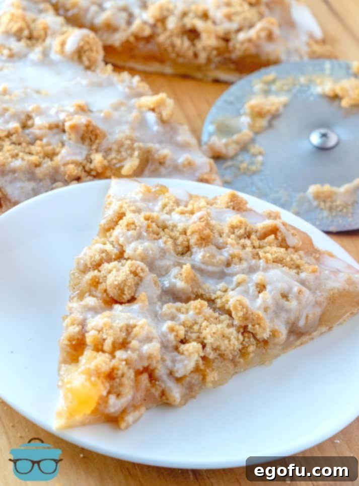 A delicious slice of Apple Dessert Pizza, topped with golden crumble and a sweet icing drizzle, rests on a small white plate, ready to be eaten.