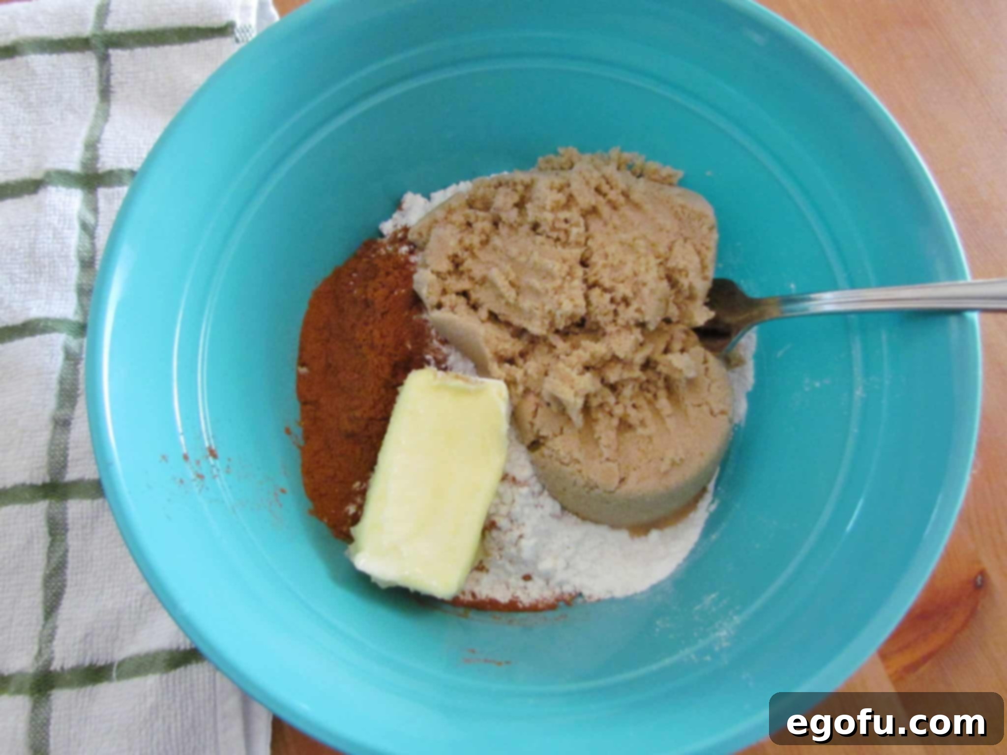 A blue mixing bowl holds the ingredients for the crumb topping: softened butter, light brown sugar, all-purpose flour, and ground cinnamon, with a fork poised to mix.