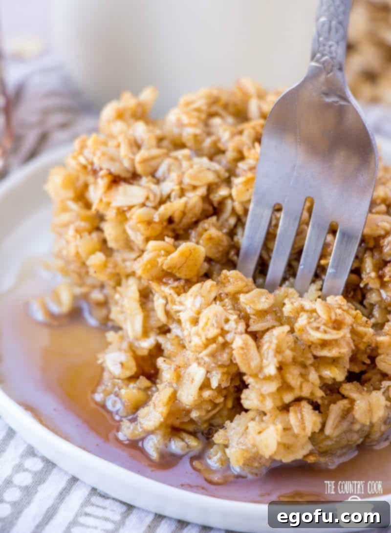 A slice of warm Maple and Brown Sugar Baked Oatmeal on a white plate, drizzled generously with maple syrup, with a fork ready to dig in. Perfect for a cozy breakfast or brunch.