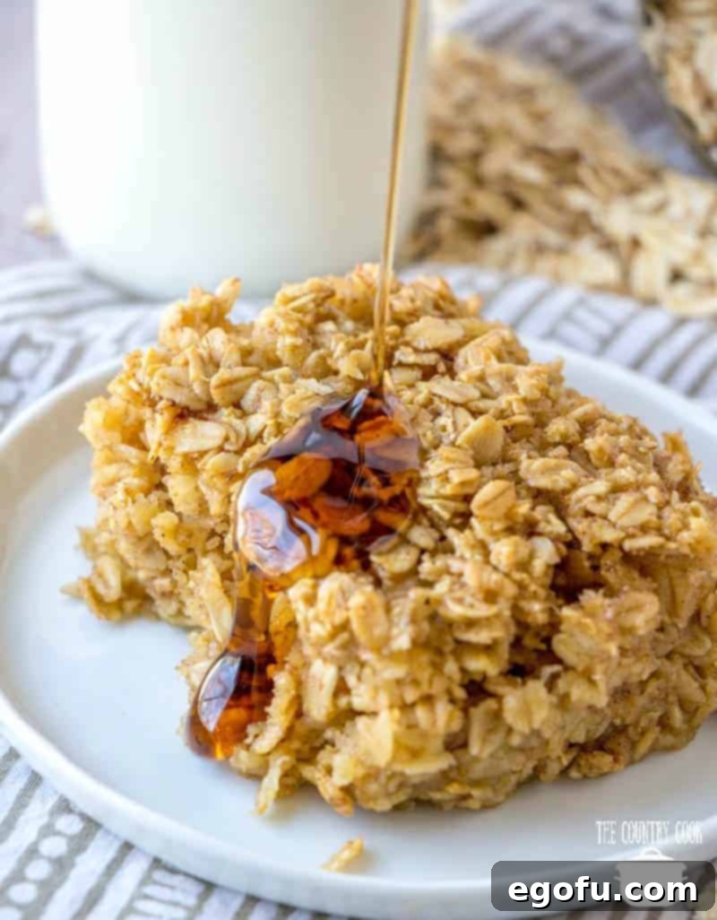A delectable slice of Maple Brown Sugar Baked Oatmeal on a round white plate, generously coated with a glistening stream of warm maple syrup, ready to be devoured.