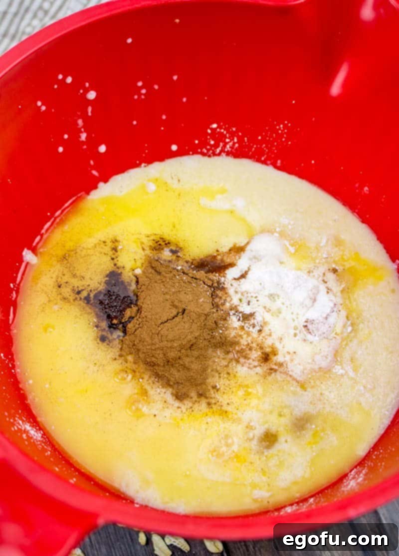 A large red mixing bowl filled with the wet ingredients for baked oatmeal: milk, brown sugar, cinnamon, beaten eggs, baking powder, and maple extract, ready to be whisked together.