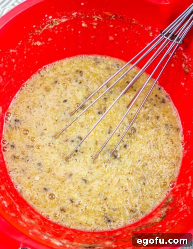 A hand vigorously whisking together the wet ingredients in a vibrant red mixing bowl, creating a smooth, cohesive batter for the baked oatmeal.
