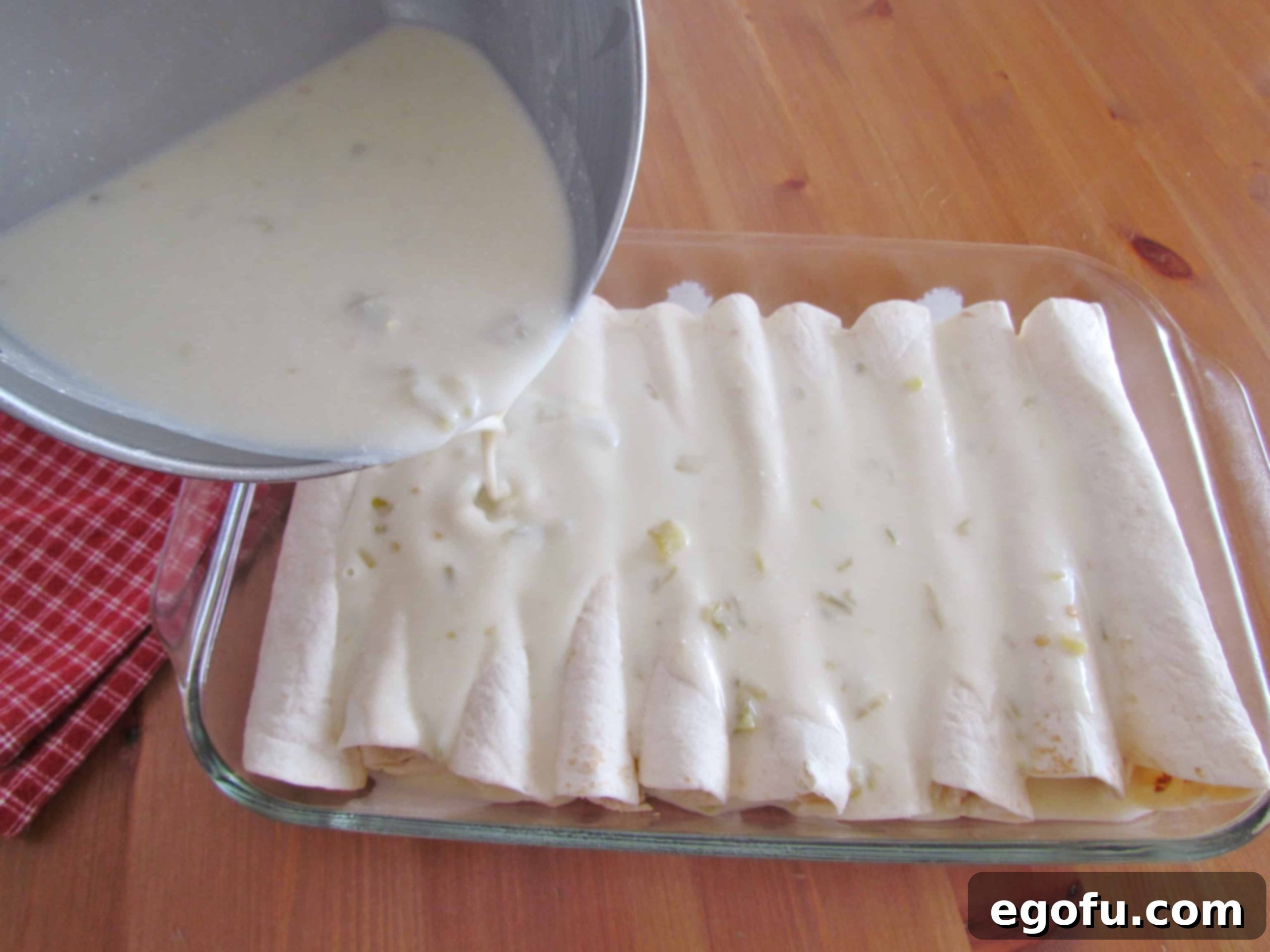 Decadent White Chicken Enchiladas 11 Pouring white sauce over chicken filled flour tortillas in a dish.