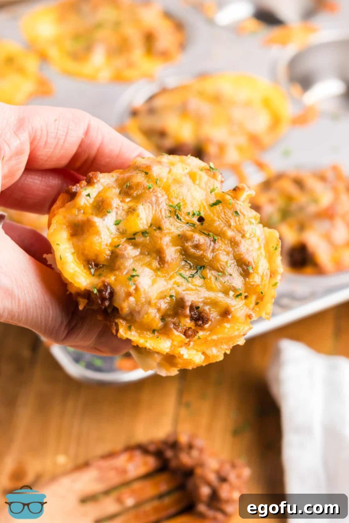 Smoky Beef Biscuit Pockets 2 A hand holding up a freshly baked BBQ Beef Biscuit Cup, showcasing its golden crust and melted cheese, over a muffin tin filled with more cups.