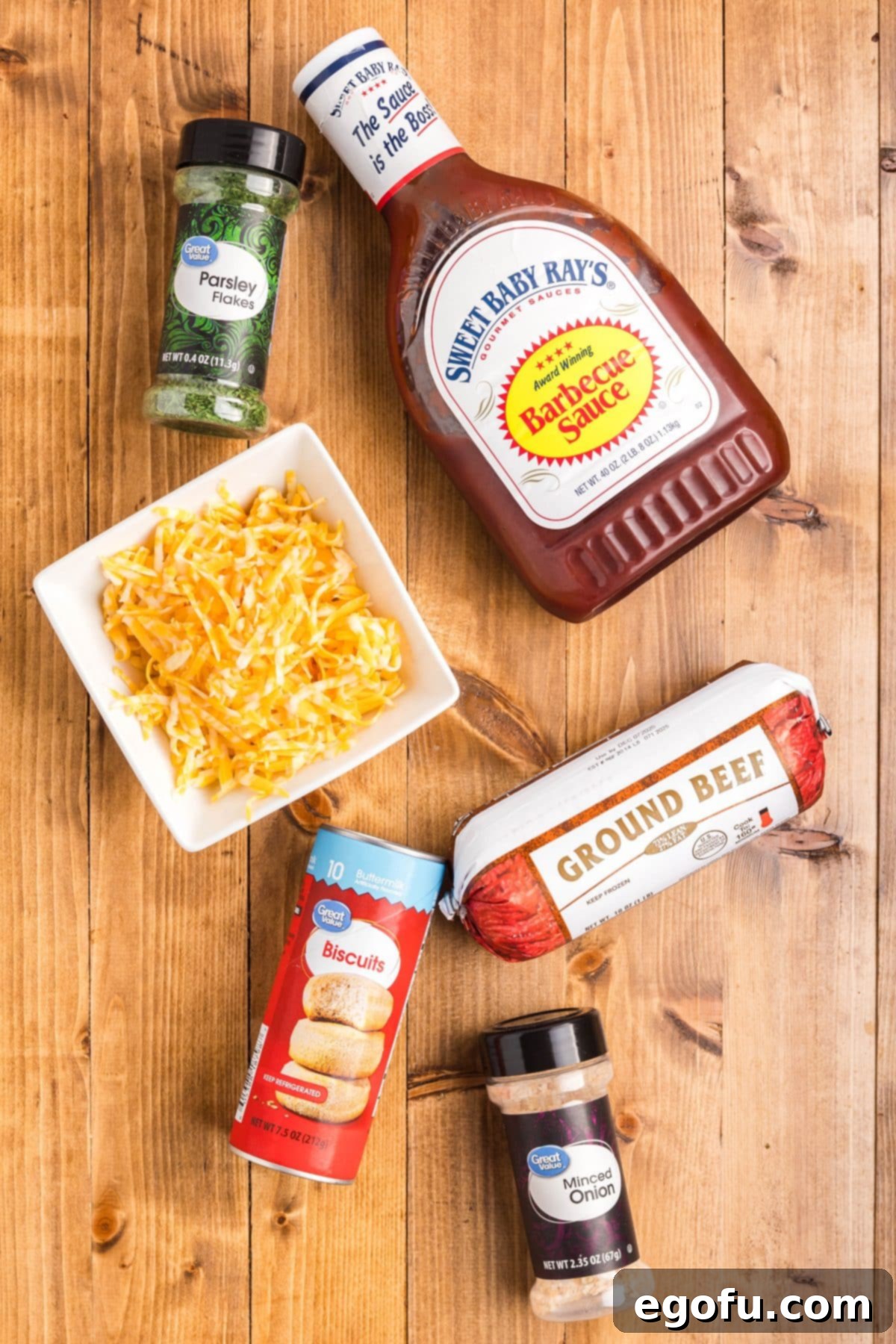 Smoky Beef Biscuit Pockets 5 A flat lay image displaying all the raw ingredients needed for BBQ Beef Biscuit Cups: ground beef, refrigerated biscuits, dried minced onion, fresh parsley for garnish, a bowl of shredded cheddar cheese, and a bottle of barbecue sauce.